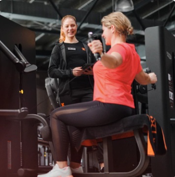 Una mujer se entrena en una estaci n de Kinesis, mientras una entrenadora FitX le gu a la ejecuci n de la rutina.