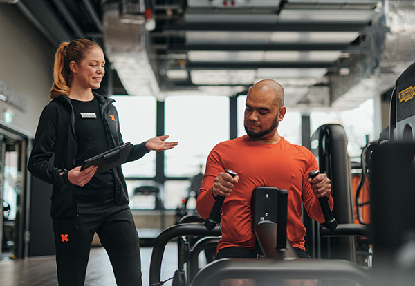 Bild: Ein Mann trainiert an einem Gerät im Gerätebereich. Neben ihm steht eine junge FitX-Trainerin mit einem Tablet und erklärt ihm die Übung.